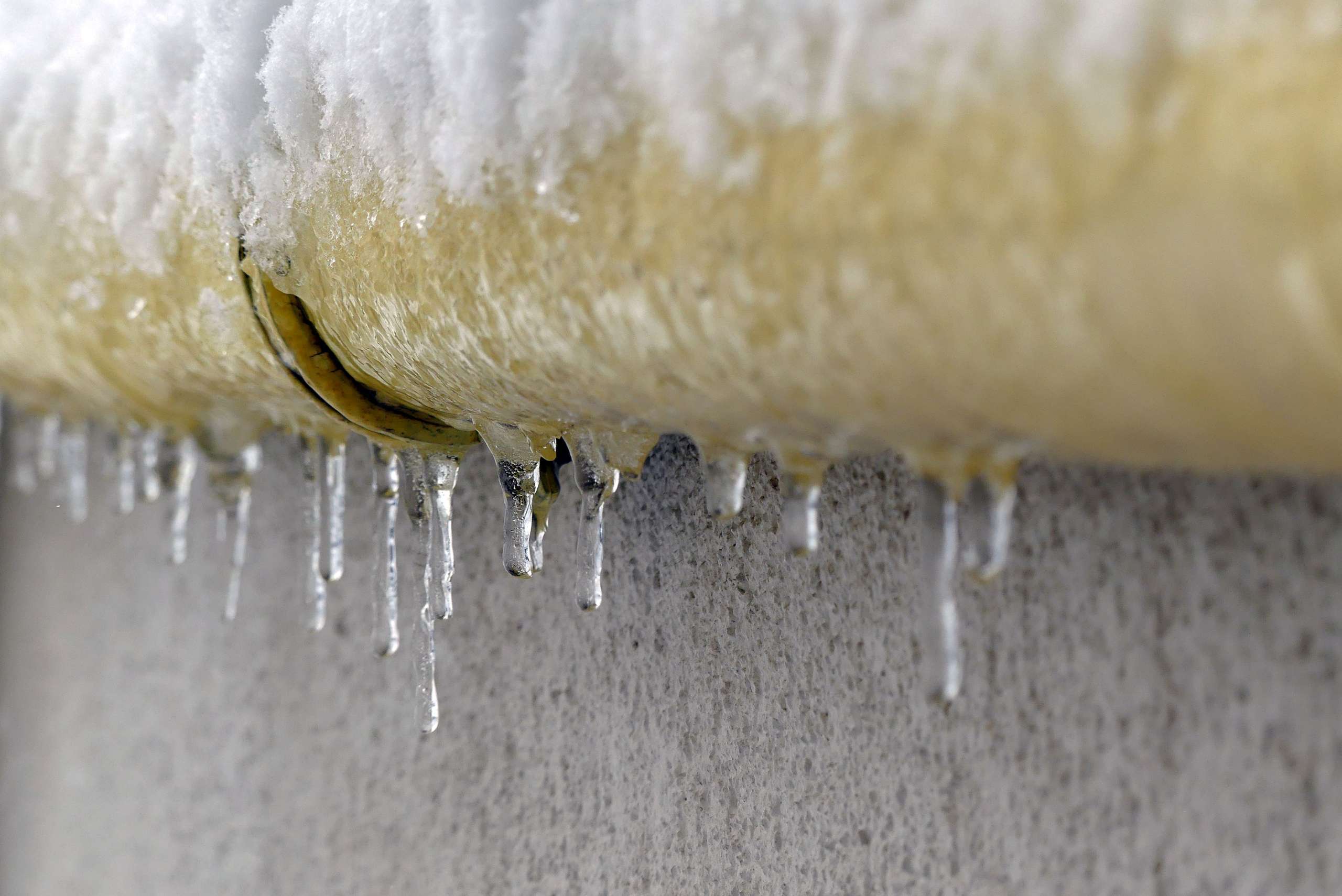 A yellow pipe on a cement wall, covered in ice and ice sickles